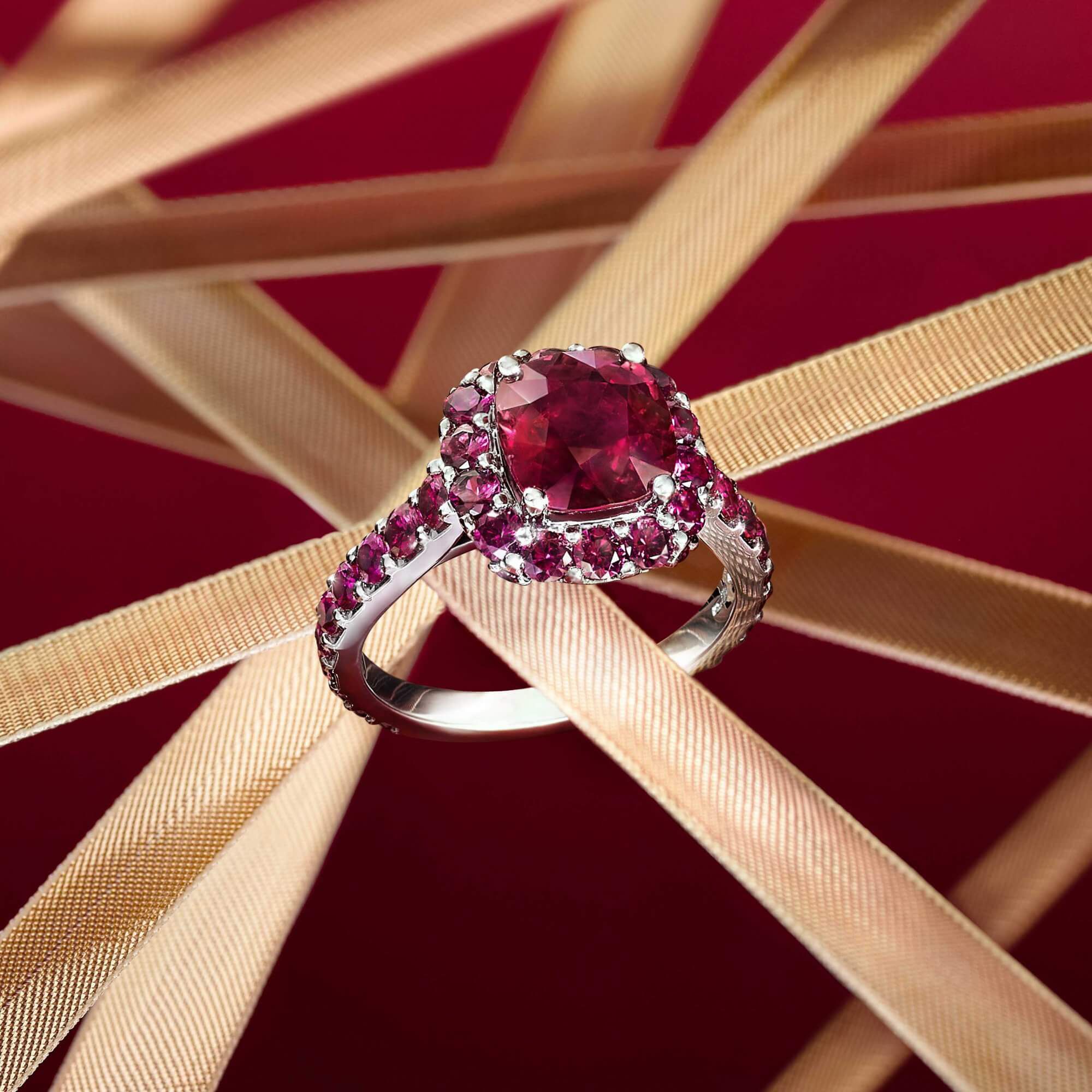 A Graff Ruby Ring featuring a cushion cut ruby with pavé round ruby surround on a red background with gold ribbons