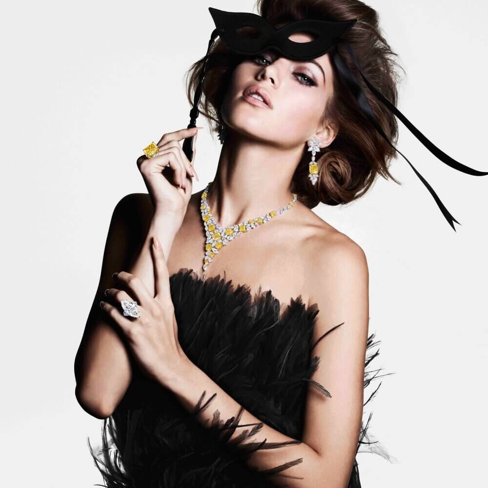 A model wearing Graff yellow and white diamond high jewellery earrings ring and necklace and a diamond ring in a black gown holding a mask