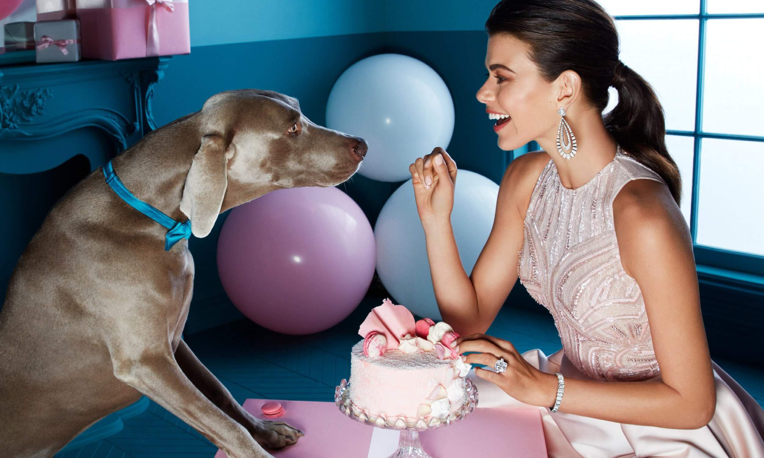A lady wearing an embroidered pink dress with baguette cut diamond earrings, an emerald cut diamond bracelet and a 20.88 carat D Flawless emerald cut diamond ring sits opposite a dog wearing a blue bow tie