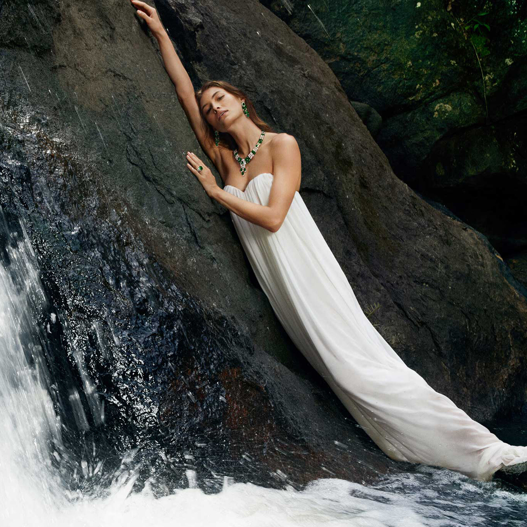 Model wears emerald and white diamond jewellery standing in waterfall