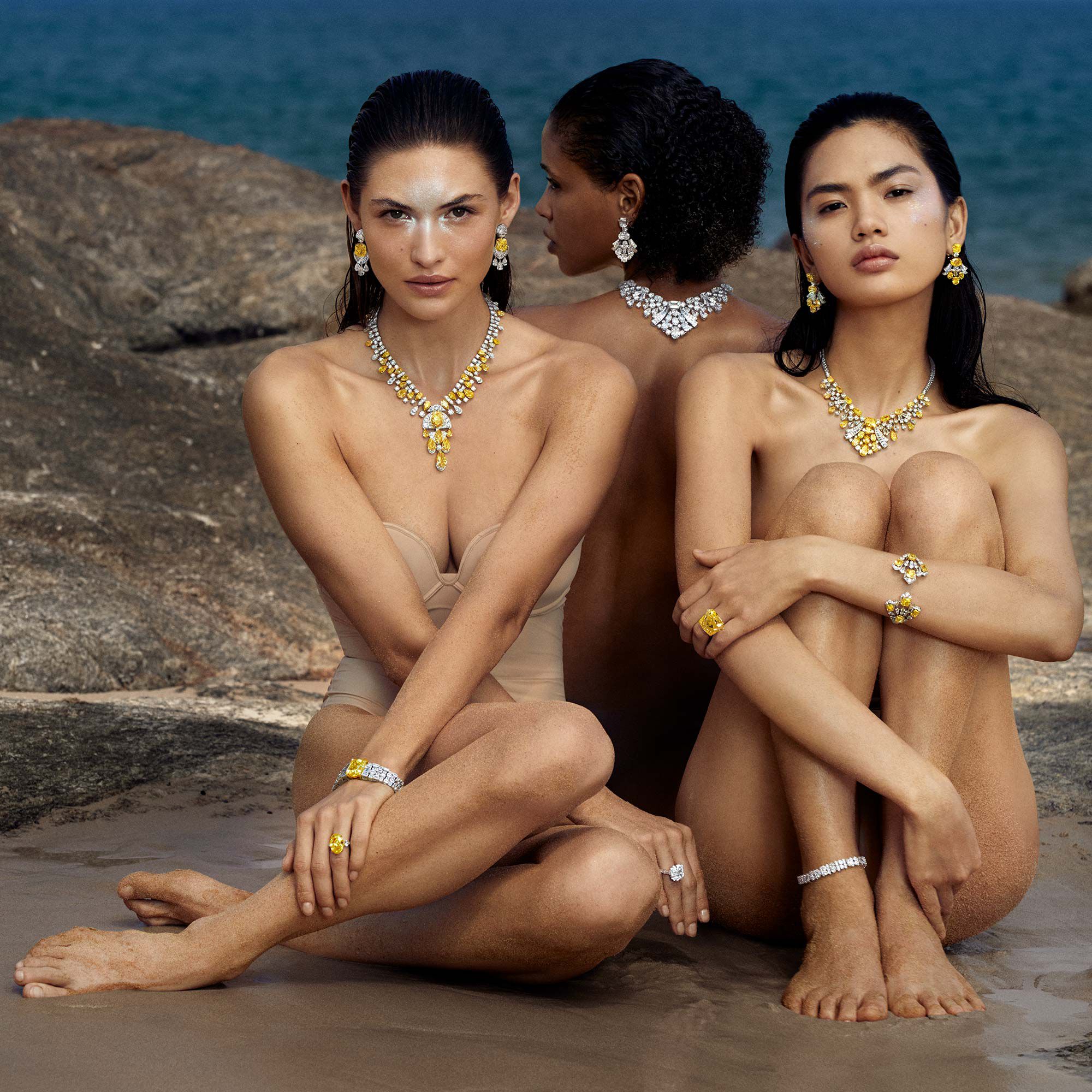 Three models wearing Graff yellow and white diamond high jewellery necklaces, earrings, rings, bracelets and ankle jewellery on a beach