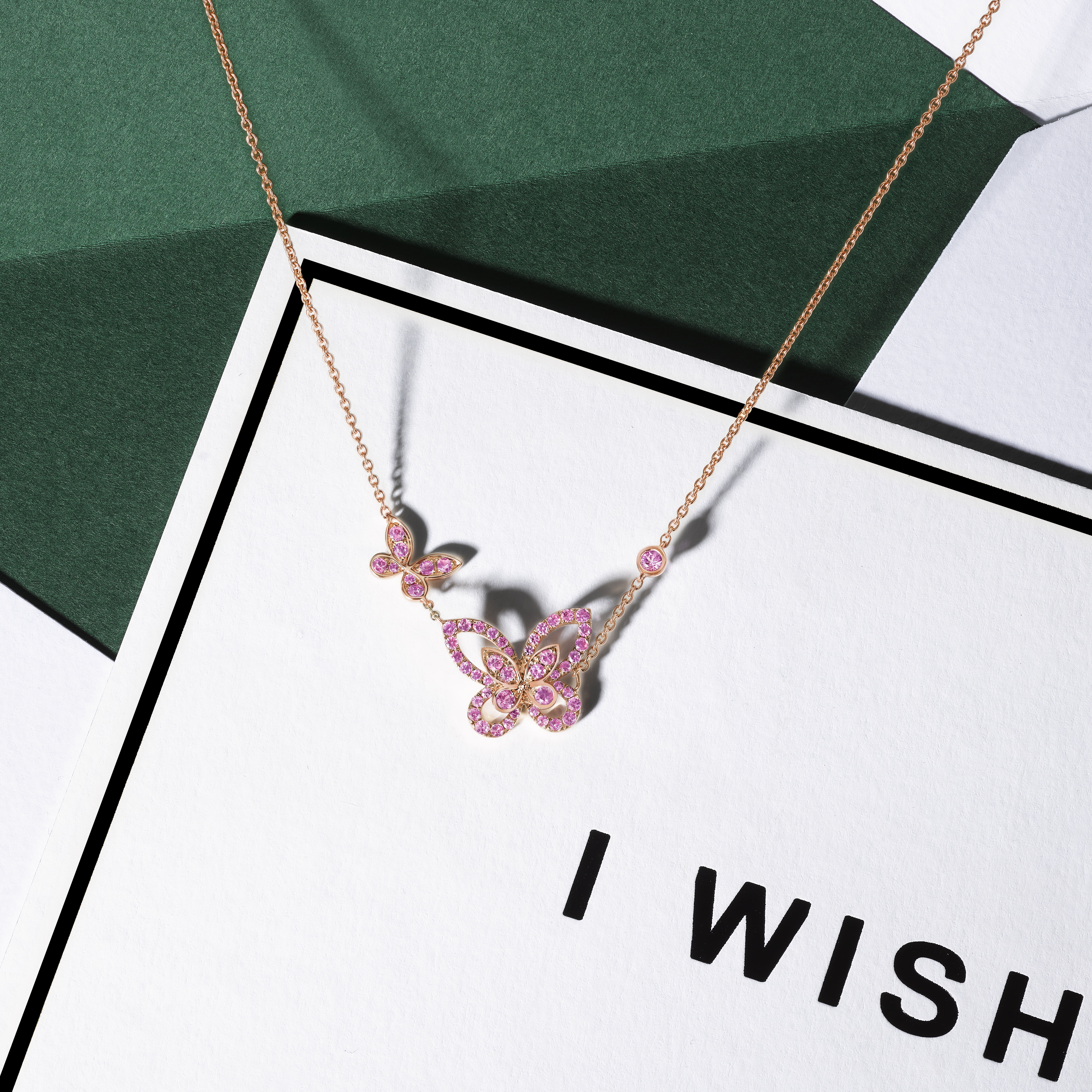 Still life image of Graff diamond collection Rose Gold and Pink Sapphire Butterfly Silhouette necklace positioned on an 'I WISH' festive greetings card