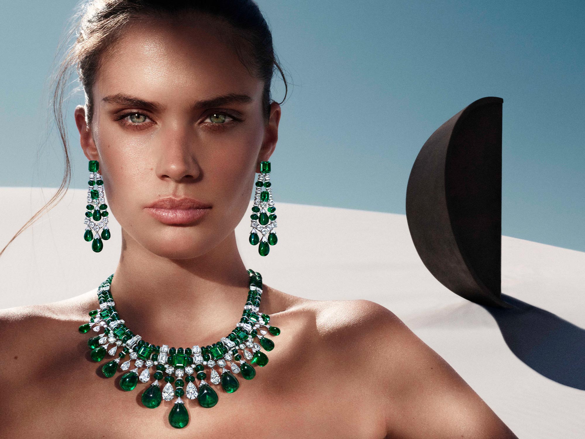 Close up of a model wearing Graff Night Moon emerald and diamond high jewellery earrings and necklace from the Tribal jewellery collection, in a desert in front of a black sculpture
